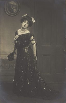 A black-and-white 20th century photograph, depicting a man in drag. He is posing is elaborate drag garb. Image was found on the "welcome collection" site of the public domain.